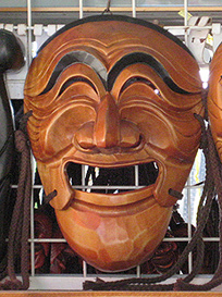 A Talchum mask in a shop in South Korea, of a laughing man with prominent eyebrows and eyes squinched shut. It is medium-brown wood with black eyebrows and a suggestion of black hair at the very top.