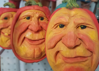 Three faces carved in wood and painted to look as though they're carved into a real pumpkin's flesh.  Each is an oval with orange 'rind' around the edges and a green pumpkin stem at the top. The faces are painted golden yellow with peach highlights on cheeks, noses and chins. The faces are of jowly elderly men with very realistic eyes.