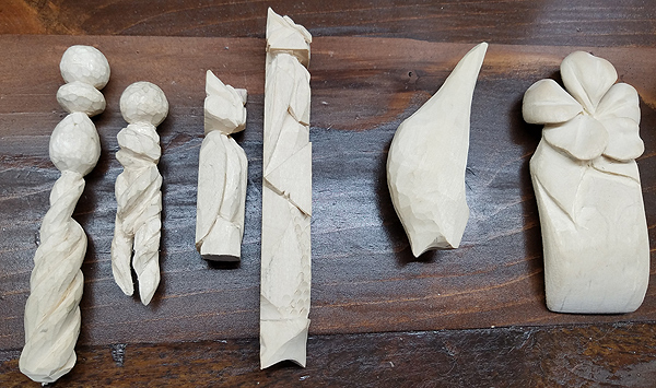 Samples of Aron's early carvings.  At left are two pieces 4-8 inches long, combining round, oval, and spiral shapes. Next, an angular abstract owl, and two even more abstract shapes. At right, a rounded block maybe 2 inches wide and six inches long, with a large tropical flower at its top