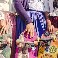 A portion of a photo showing three women wearing polleras, standing close together, each holding a skateboard by its end.