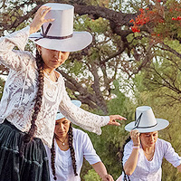 A portion of a photo showing three women riding skateboards and wearing tall white bowler hats. They each have a hand on their hat to keep it in place.