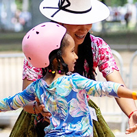 A portion of a photo showing a little girl in a pink helmet learning to ride a skateboard. A member of ImillaSkate stands behind her, one hand on the little girl's back to guide her. The little girl is holding her arms out to her sides and looks excited and happy.