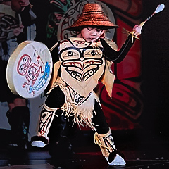 A small child demonstrates a Tsimshian dance, a tradition of indigenous people of the Pacific Northwest. He wears a tunic decorated with a stylized face, matching leggings, and a straw hat with angled sides. He holds a large flat drum out to his right and a drum-beater out to his left.  He is looking down.  His feet are wide apart, and one foot is raised.  The drum is about 14 inches wide and three inches deep, and its skin head is decorated with a stylized figure holding a spear.