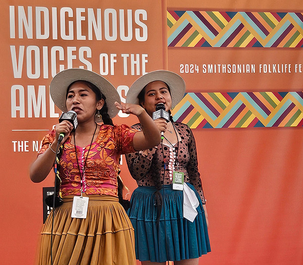 Two young women from Bolivia are performing hip-hop at the festival. Their garments echo traditional Bolivian garb, with a contemporary twist: colorful bulky skirts with the hem at mid-thigh, close-fitting tops with deep necklines and lots of buttons down the front, and straw hats with wide brims. Their black hair is in long braids. They appear to be in their 20s.