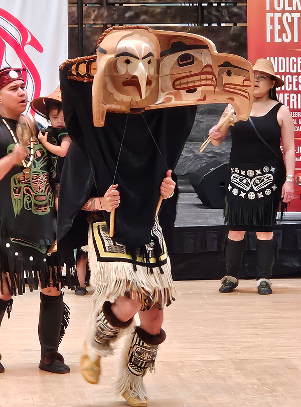 A member of the Tsimshian people performs a traditonal dance, wearing a large stylized eagle head mask which appears to be about three feet wide. A black drape covers the dancer’s upper body, with fringed boots and knee-length skirt completing the costume.  Two musicians stand behind the eagle figure, dressed in black clothing with stylized decorations. The nearer musician, a man, is holding a toddler dressed in the same clothing.