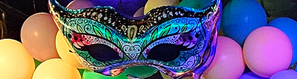 A large mylar balloon in the shape of a mask that would cover a person's eyes. It features jewel-tone graphics against black. It is displayed in front of a mass of balloons in similar colors.