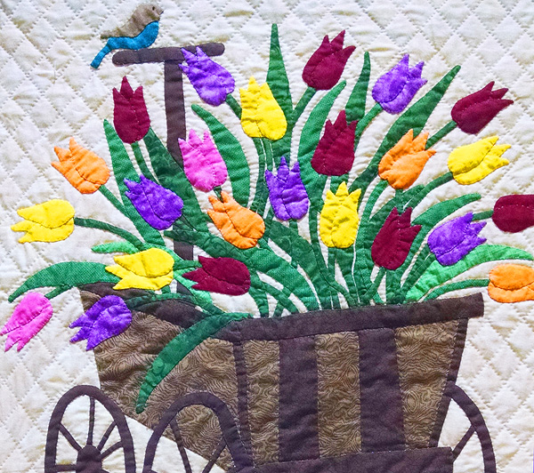 An applique quilt showing a colorful array of tulips in a wooden cart or buggy. A bluebird perches on the cart’s handle.