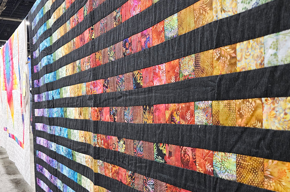 Quilts are displayed hanging over a long clothes-line. The one in the foreground is alternating horizontal strips of black and bright colors. The colors are selected to create arcs of color centered on the far edge of the quilt, shadingfrom lavendar to blue to yellow to red.