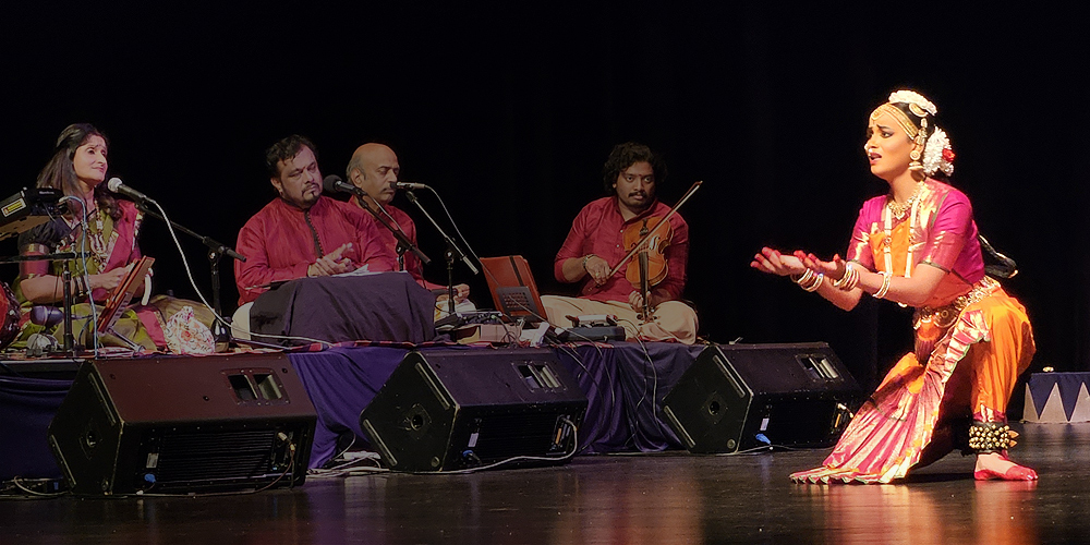 A young woman performs a classical Indian dance.She is crouching with her arms out in front of her as if she holds a box, and she looks worried. At left are four musicians sitting cross-legged on a low platform. The dancer's dress is magenta and orange, with a pleated skirt, and decorations on her head.