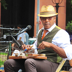 A Black man plays a washboard on his lap, peering out from under a straw fedora hat. He is wearing a white shirt, a red tie, and a grey vest and slacks without the suit jacket.