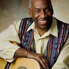 A Black man is smiling warmly, resting his hand on the upper curve of a guitar.  He has short graying hair, a wide smile, and a long face. He wears a white shirt and a vest with narrow vertical ribbon-like stripes.